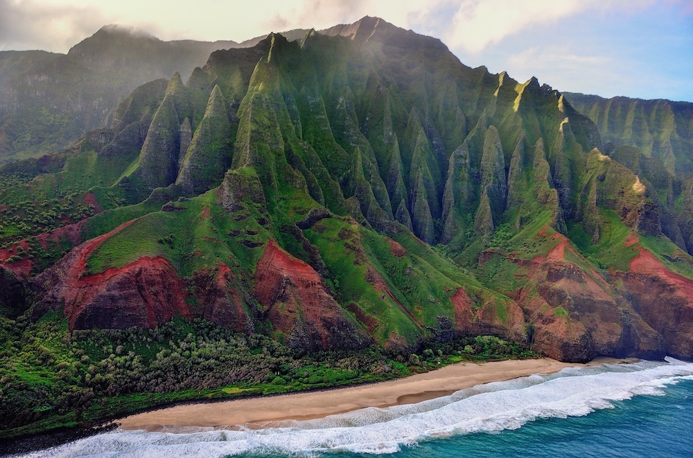 Tall mountains with red and green along the Napali Coast in Kauai with the ocean waves rolling in on shore