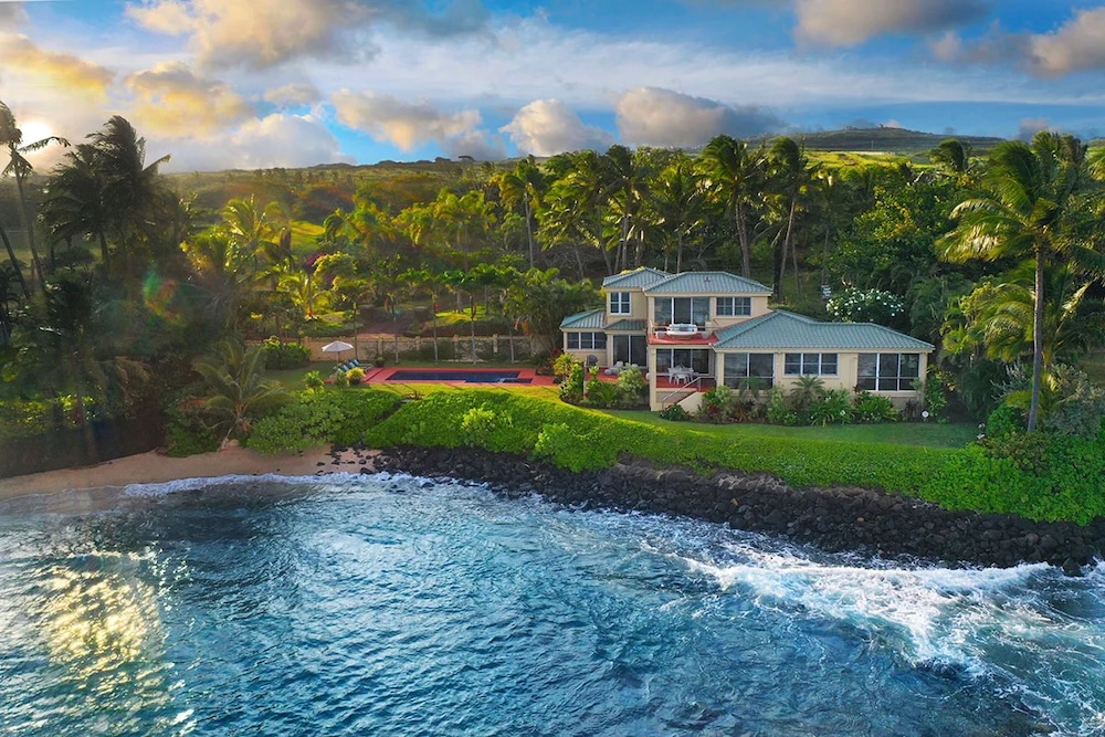 beach house on the shore in kauai with waves rolling in and the green palm trees and mountains in background